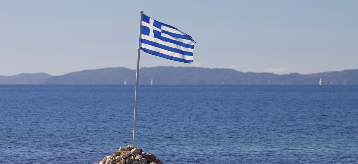 Greek flag waves over rocky island with serene Ionian Sea backdrop in Corfu, Greece.