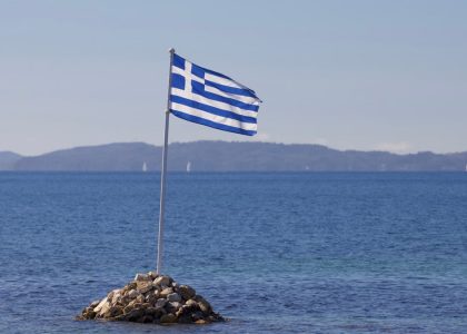 Greek flag waves over rocky island with serene Ionian Sea backdrop in Corfu, Greece.